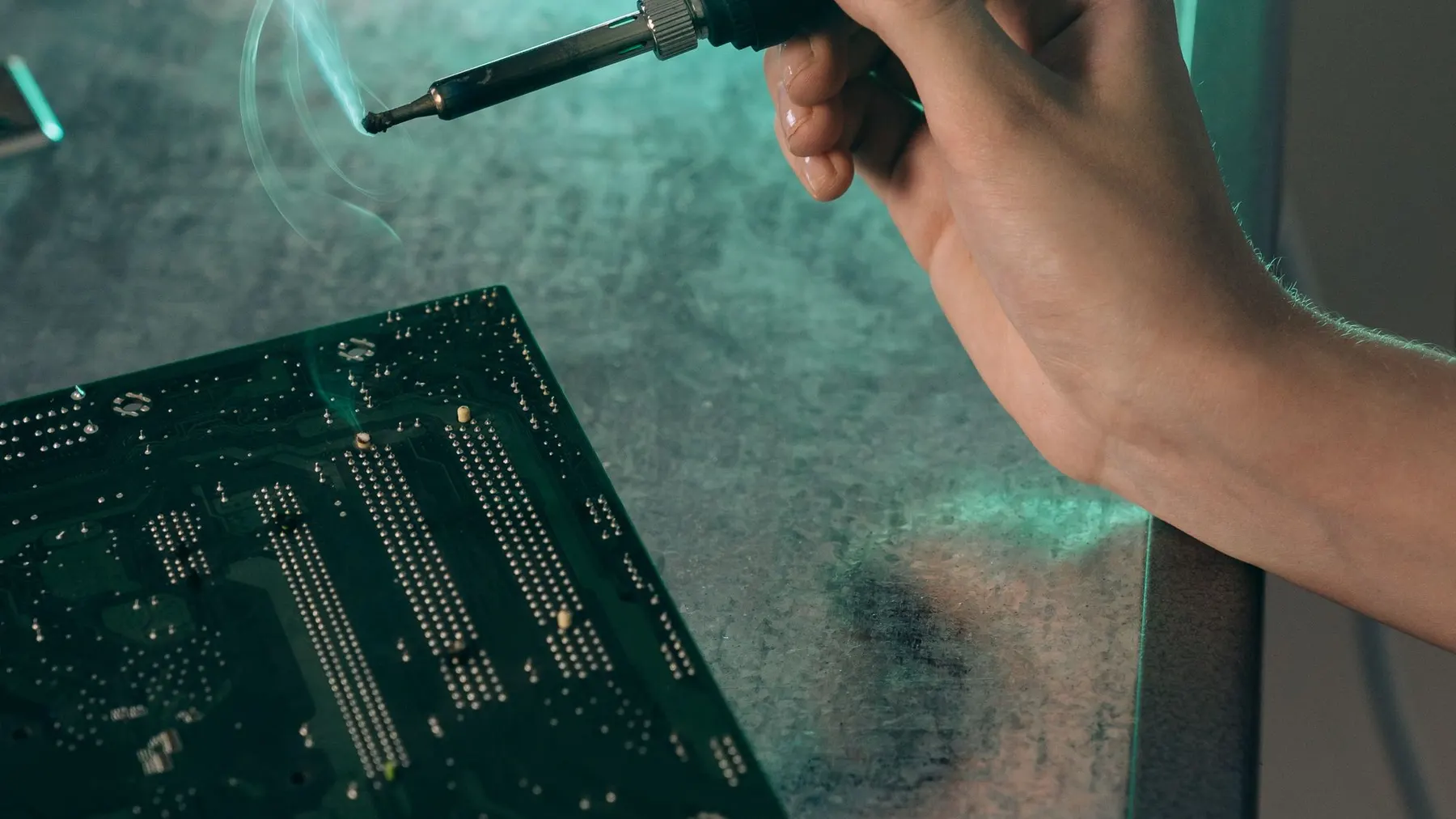 Fer à souder en main au-dessus d’une carte mère, la fumée du flux encore visible.
