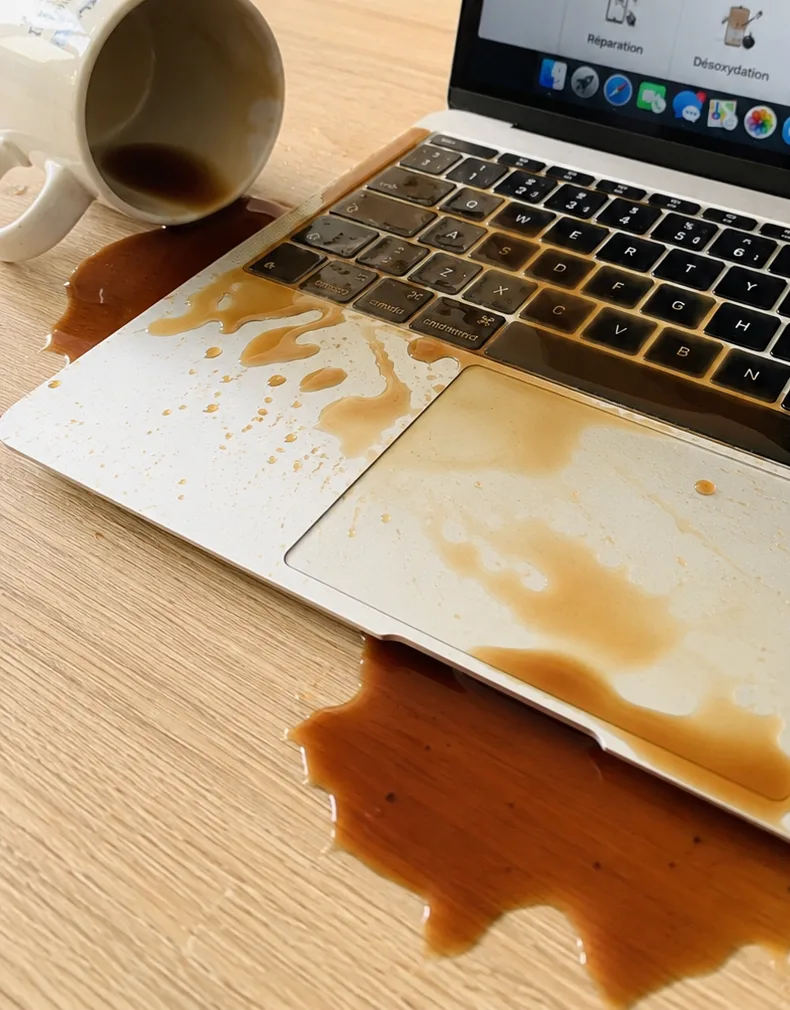 Une tasse renversée sur un MacBook : le café a coulé entre les touches, sur le trackpad et jusque sur le bureau.